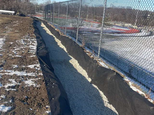 Finished French drain, fabric-lined and stone-filled, along a ballfield fence