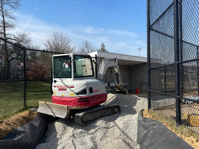 Mini excavator placing stone in a fabric-lined trench at a ballfield